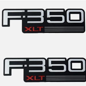 Puede incluir: Dos emblemas negros y plateados F350 XLT. Los emblemas tienen forma rectangular con el texto "F350" en blanco y "XLT" en rojo. Los emblemas tienen un borde plateado y están sobre un fondo blanco.