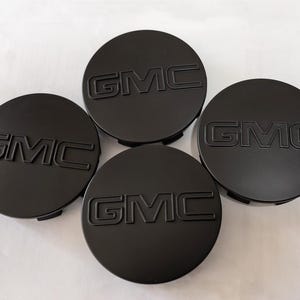 May include: Four black GMC center caps arranged on a white surface. Each cap is circular with the embossed letters "GMC" in the center. The caps are a matte black finish, and the logo is raised.