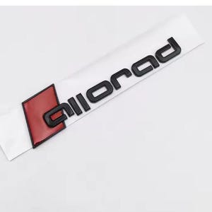 May include: A rectangular automotive emblem featuring the word allroad in raised black lettering. To the left of the text is a red parallelogram shape with a black border. The entire emblem is mounted on a white adhesive backing strip.