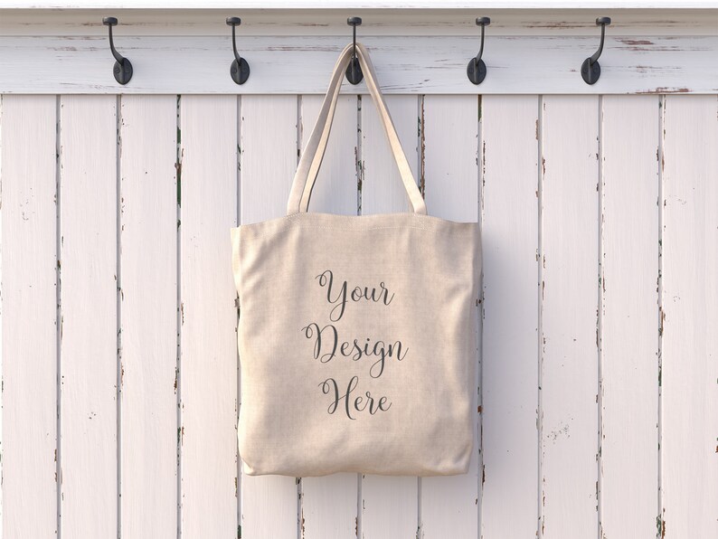 May include: A beige canvas tote bag hanging on a white wooden rack with hooks. The bag has the text "Your Design Here" printed on the front.