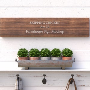 May include: A farmhouse style wooden sign mockup with the text "Skipping Cricket 6 x 28 Farmhouse Sign Mockup". The sign is hanging on a white shiplap wall with a shelf below. The shelf has a metal planter with five small green plants and three hooks. Two of the hooks have a brown leather strap and a plaid scarf hanging from them.