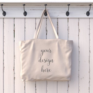 Op de afbeelding: Een effen witte canvas draagtas die aan een witte houten hakenrek hangt. De tas heeft de tekst "your design here" op de voorkant gedrukt.