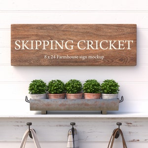 Könnte beinhalten: Ein Holzschild mit dem Text "SKIPPING CRICKET" in weißen Buchstaben. Das Schild ist 20,3 cm breit und 61 cm lang und ist ein Farmhouse-Schild-Mockup. Das Schild hängt an einer weißen Wand mit einem Regal darunter. Das Regal hat einen Metalltopf mit fünf kleinen grünen Pflanzen darin. Es gibt drei Haken am Regal.