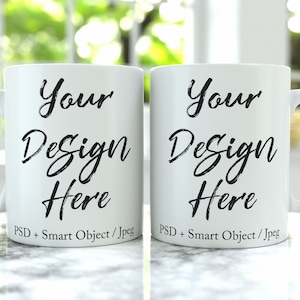 以下が含まれることがあります： 白いセラミック製のマグカップ2つ。黒い文字で「Your Design Here」と「PSD + Smart Object / Jpeg」と書いてあります。