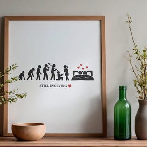 Puede incluir: Impresión enmarcada con un diseño humorístico sobre la evolución del amor. Las figuras de silueta negra representan etapas desde las primeras formas humanas hasta una pareja en la cama. El texto "STILL EVOLVING" está debajo, con un corazón rojo.