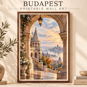 Puede incluir: Una impresión de arte mural enmarcada titulada BUDAPEST PRINTABLE WALL ART que muestra una vista a través de un arco de piedra hacia un castillo con torres, un río y el horizonte de la ciudad al atardecer. Un farol ilumina la terraza de piedra.