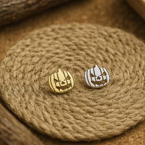 May include: Two small pumpkin-shaped pins. One pin is gold-colored, and the other is silver-colored. Both pins have a carved jack-o'-lantern face design. They are displayed on a round, woven, tan-colored mat.