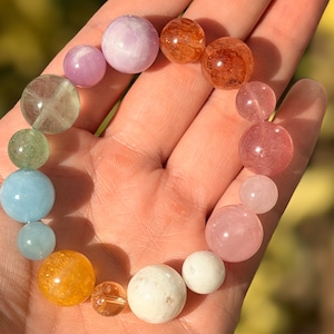 May include: A colourful beaded bracelet with a variety of gemstones. The bracelet features round beads in shades of pink, orange, green, blue, and white. The bracelet is held in a hand, with a blurred background.