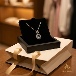 May include: A silver necklace featuring a large, oval, pale blue gemstone pendant, encircled by smaller clear stones. The necklace is presented in a black velvet box, on top of a white gift bag with a gold ribbon. The brand name "CLAIRORA" is visible.