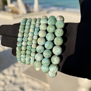 May include: Several turquoise and white beaded bracelets. The bracelets feature round and square beads of different sizes. They are presented on a black surface, with a beach and ocean visible in the background.
