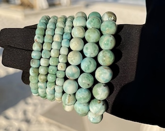 Könnte beinhalten: Eine Sammlung türkisfarbener und weißer Armbänder mit Perlen. Die Armbänder bestehen aus runden und quadratischen Perlen in verschiedenen Größen. Sie werden auf einer schwarzen Oberfläche präsentiert, mit Strand und Meer im Hintergrund.