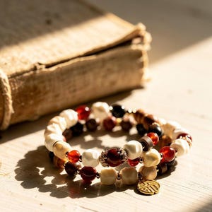 May include: Three beaded bracelets featuring cream, brown, red, and gold beads. The bracelets are stacked on a light wooden surface, with a burlap-wrapped object in the background. A gold charm hangs from one bracelet.