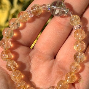 May include: A bracelet made of golden-yellow citrine gemstone beads. The bracelet has a clear, larger bead with a central rod and small, sparkling accents. The beads are strung together, creating a piece of jewellery.