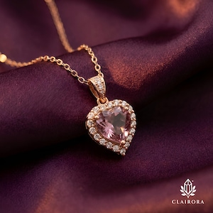 May include: A gold-tone necklace with a heart-shaped pendant. The pendant has a pink gemstone centre, encircled by small, clear stones. The necklace is displayed on a deep purple, textured fabric. The brand name "CLAIRORA" is visible.