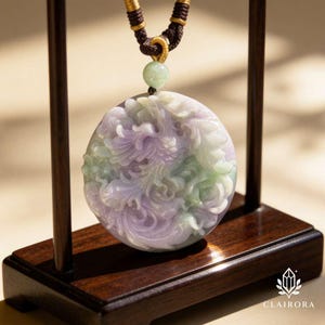 May include: A round jade pendant with intricate carvings in shades of lavender, green, and white. The pendant is suspended from a brown and gold cord. The pendant is displayed on a wooden stand.