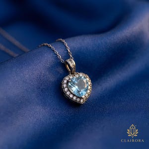 May include: A silver necklace with a heart-shaped pendant. The pendant has a light blue gemstone surrounded by small, clear stones. The necklace is displayed on blue satin fabric. The brand name "CLAIRORA" is visible in the bottom right corner.
