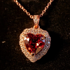 May include: Rose gold necklace with a heart-shaped pendant. The pendant has a large, faceted red gemstone surrounded by a halo of smaller, clear gemstones. The chain is delicate and rose gold.