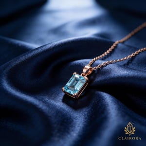 May include: A gold necklace featuring a rectangular, pale blue gemstone pendant. The pendant is set in a gold frame and suspended from a fine chain. The necklace is displayed on a dark blue satin fabric, creating a luxurious aesthetic.