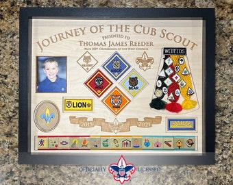 May include: A framed wooden display commemorating a Cub Scout's journey. It features a photo, badges, and the text "Journey of the Cub Scout." The display includes badges for Wolf, Bear, Tiger, and Lion, along with a Webelos rank. The frame is black.