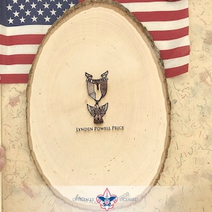 May include: A wooden round plaque with a brown Eagle Scout award displayed on it. The plaque has the text "Lynden Powell Price" engraved on it. The plaque is in front of an American flag.