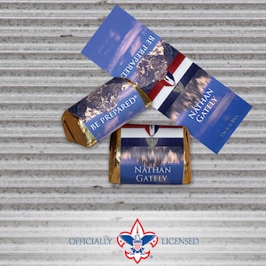 May include: Three chocolate bars with gold foil wrappers and blue labels. The bars have the text "BE PREPARED" and "NATHAN GATELY". The image also includes a blue and white card with the text "BE PREPARED" and "NATHAN GATELY". The official Boy Scouts of America logo is at the bottom.