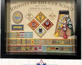 May include: A framed Cub Scout achievement display. The wooden background features patches and awards, including badges for Lion, Tiger, Bear, and Wolf. The frame includes the text "Journey of the Cub Scout" and "Presented to John R. Chavez."