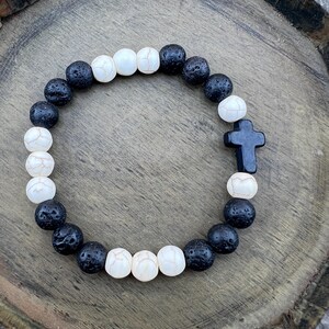 May include: A bracelet made of black lava rock beads and white, textured beads. A black cross charm is featured in the centre. The bracelet is displayed on a wooden surface.