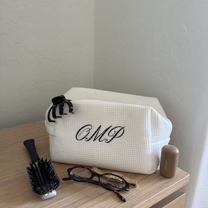 May include: A white, textured cosmetic bag with the embroidered initials "OMP" is displayed on a wooden surface. A black hair clip, a round hairbrush, eyeglasses, and a small brown container are also present.