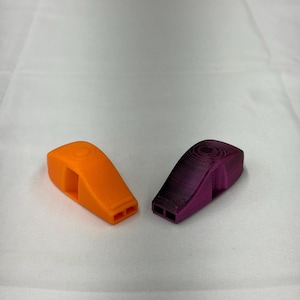 May include: Two plastic whistles, one orange and one purple, are displayed on a white surface. The whistles have a rectangular shape with two holes at the bottom and a circular design on top.