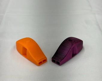 Op de afbeelding: Twee plastic fluitjes, één oranje en één paars, worden weergegeven op een wit oppervlak. De fluitjes hebben een rechthoekige vorm met twee gaten aan de onderkant en een cirkelvormig ontwerp aan de bovenkant.
