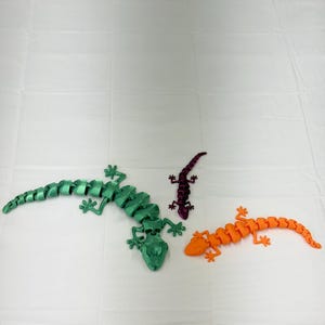 Peut inclure: Trois figurines de lézard articulées en vert, orange et violet. Les lézards sont faits de pièces segmentées, permettant une pose flexible. Le lézard vert est le plus grand, suivi des lézards orange et violet. Le fond est une surface blanche unie.