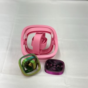 May include: A collection of 3D-printed geometric sculptures in shades of pink, green, and purple. The largest sculpture is a pink square with an interior design. The other two are smaller, one green and gold, the other purple and black.