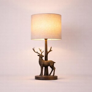 May include: A decorative table lamp with a bronze-coloured deer figurine and antlers, positioned beneath a tree-like support. The lamp has a beige fabric lampshade. The base is round. The lamp is set against a neutral background.