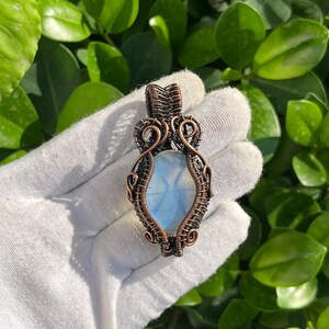 May include: A handcrafted pendant with a light blue, oval gemstone, set within detailed copper wire. The wirework has an intricate, woven pattern, creating a unique, artistic piece. The pendant is shown against a backdrop of green foliage.