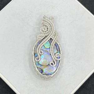 May include: A handcrafted pendant featuring an iridescent abalone shell encased in intricate silver wire. The wirework forms a decorative frame and bail, showcasing the shell's natural colors of blue, green, and gold. This unique jewelry piece is designed to be worn as a necklace.