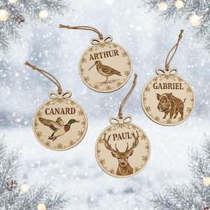 Peut inclure: Quatre décorations de Noël en bois avec des illustrations d'animaux gravées et des noms : Arthur, Canard, Gabriel et Paul. Chaque ornement a une boucle en ficelle pour l'accrocher et des détails de flocons de neige. Les ornements sont circulaires et en bois clair.
