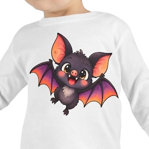 May include: A white long-sleeve shirt featuring a graphic of a small, dark purple bat with large eyes, a pink nose, and wings that transition from purple to orange. The bat has a smiling expression with visible white teeth and a light-colored chest.