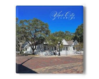 May include: A square canvas print featuring a photograph of a historic white building with a porch, trees, and a brick road. The text "Ybor City FLORIDA" is in the upper right corner against a bright blue sky.
