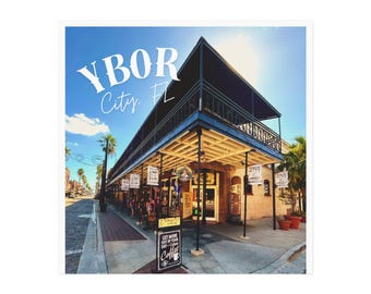 May include: An exterior shot of a two-story building with a balcony, awnings, and a sign that reads "Ybor City, FL." The building is made of brick and has several signs hanging from the awnings. A sign in front advertises coffee.
