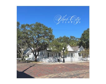 May include: A street scene featuring white buildings with porches and a red brick road. Lush green trees frame the buildings under a clear blue sky. The text "Ybor City FLORIDA" is displayed in an elegant script across the top.