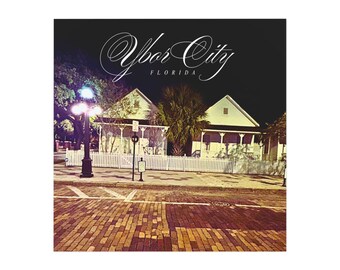 May include: A nighttime street scene featuring white houses behind a picket fence. The image has a vintage feel, with the text "Ybor City Florida" in elegant script across the top. The street is paved with bricks, and a lamppost illuminates the scene.