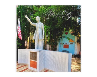 May include: A white statue of a man in a suit with his arm raised, set on a white tiled base. The image includes an American flag and a sign that says "Ybor City Florida". A mural with the words "Isla de Cuba" is in the background.