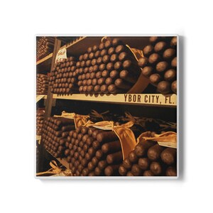 May include: A square print featuring stacks of cigars on wooden shelves. The cigars are brown and neatly arranged, with the text "YBOR CITY, FL." visible. The image has a vintage, sepia-toned aesthetic.