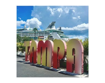 May include: Large, colorful sign spelling "TAMPA" with a gradient from yellow to red. A cruise ship is in the background under a blue sky with fluffy white clouds. Palm trees and greenery are visible.