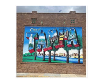 May include: A large mural on a brick building spells out "TAMPA" in 3D letters. The mural depicts scenes of Tampa, Florida, including a lighthouse, palm trees, and a river. The words "City of" are above the "T".