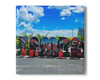 May include: A square canvas print displaying the word "TAMPA" in large, colourful letters. Each letter is decorated with images of the Tampa Bay Buccaneers football team, including their logo and team colours. The background is a bright blue sky with fluffy white clouds.