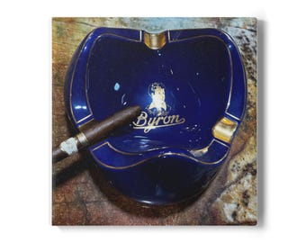 May include: A navy blue ashtray with gold trim and the name "Byron" in gold lettering. A lit cigar rests in the ashtray. The ashtray has a heart-shaped design.
