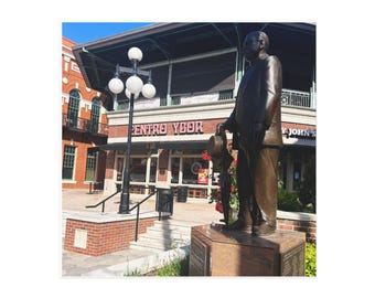 May include: A bronze statue of a man in a suit holding a hat, standing on a granite base. The statue is in front of a building with the text "CENTRO YBOR" and "Y JOHN'S". A black lamppost with white globe lights is to the left.