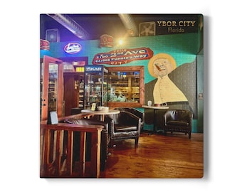 May include: Interior shot of a cigar lounge with dark leather chairs, wooden tables, and a mural on a teal brick wall. Neon signs and the text "Ybor City Florida" are visible.