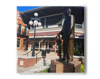 May include: A bronze statue of a man in a suit holding a hat, standing on a stone pedestal. The statue is in front of a building with the text "CENTRO YBOR" and "Y JOHN'S". A street lamp with white globe lights is to the left.